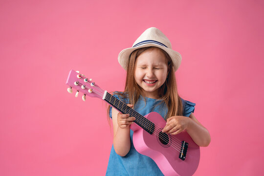 cheerful little girl in a hat plays a pink ukulele guitar.