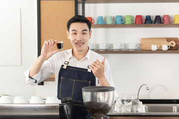 Smiling Asian barista man show cup of black coffee, thumps up feeling happy look at camera in coffee shop