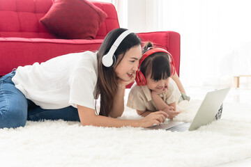 Happy Asian mother and little daughter put on the headphone and look at the screen of laptop, smile and joyful, spend time with family on holiday