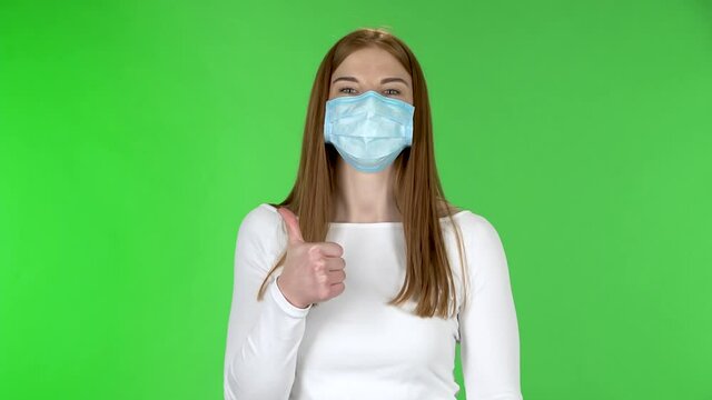 Portrait of happy pretty young girl in medical protective face mask looking at camera and showing thumbs up gesture with both hands.
