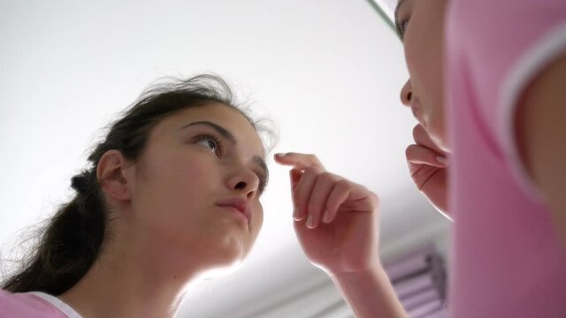 teenage brunette approaches large mirror and pops zits on forehead with fingers at home in morning close view