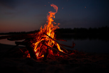 Bonfire on the river Bank at night. Red flames from burning branches and firewood near the reservoir in summer. Outdoor recreation by the fire.
