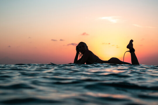 Portrait from the water of surfer girl with beautiful body on surfboard in the ocean at colourful sunset time