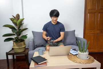 handsome asian freelancer working on his laptop computer remotely from home, sitting on the cozy beige sofa at home, wearing casual outfit.