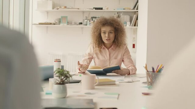 Over-the-shoulder shot of young woman with curly hair sitting at office table in front of her unrecognizable colleagues holding her day planner telling something to them