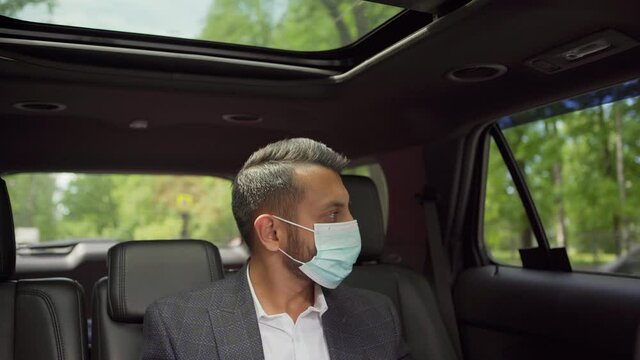 Tilt up of young Middle Eastern businessman in suit putting on face mask sitting in backseat of private car or taxi with laptop on his lap