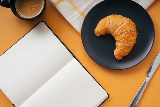 Top view - Golden crossiant on black plate with knife and hot coffee over orange background. With the book.