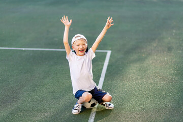 a blond boy in a cap in a sports uniform sits on a soccer ball on the football field and shouts Goal, sports section. Training of children, children's leisure