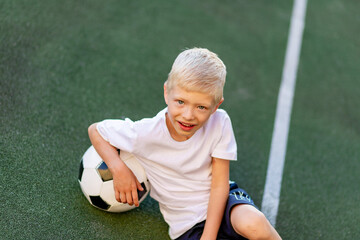 a blond boy in a sports uniform sits on a football field with a soccer ball, sports section. Training of children, children's leisure