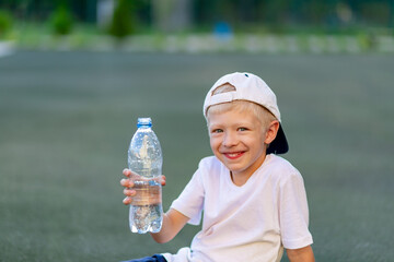 portrait of a blond boy in a sports uniform sitting on a green lawn on a football field and holding a bottle of water. Training children