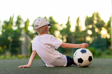 a boy in sports clothes sitting on a green lawn on a football field with a soccer ball back, back view, sports section, training