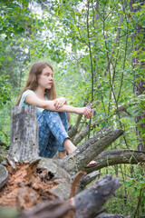 In the summer, a girl sits in a forest on a fallen tree.