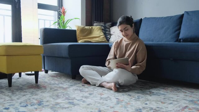 Mature Indian woman sitting Near sofa and websurfing on digital tablet