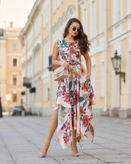 Young beautiful elegant tall slim woman with natural makeup and wavy brunete hair wearing colorful dress and sunglassses walking in the city on a summer day and holding handbag