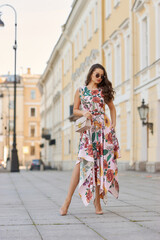 Fototapeta premium Young beautiful elegant tall slim woman with natural makeup and wavy brunete hair wearing colorful dress and sunglassses walking in the city on a summer day and holding handbag