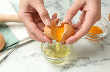 Woman separating egg yolk from white over glass bowl at marble table, closeup