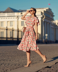 Pretty lady wearing wrap around dress. Beautiful woman with wavy brunette hair walking un the city. Elegant girl with long leegs in high heels. Fashionable female model in sunglasses holding handbag