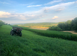 Fototapeta premium Golden hour sunlight makes colors soft and image idyllic. Off-road vehicle sits on a grassy hill atop an agricultural field landscape with copy space. High quality photo in natural light, with rural