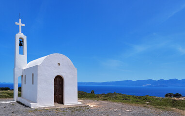 White Greek Orthodox chapel or church on hilltop of seashore against clear blue sky on sunny day. 