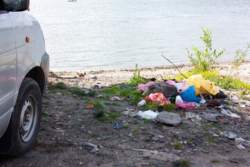 Bank of the river or lake. Car and trash near the wheel on the beach - plastic bags, paper cups, paper, glass, bottles