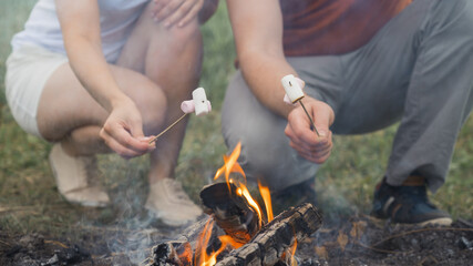 Guy and the girl are frying marshmallows on a campfire. Friends hold sticks with marshmallows over a bonfire. Horizontal orientation, selective focus.