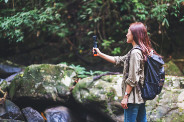 A female tourist with backpack shooting video of the waterfall in the forest by an action camera
