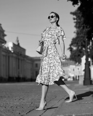 Pretty lady wearing wrap around dress. Beautiful woman with wavy brunette hair walking un the city. Elegant girl with long leegs in high heels. Fashionable female model in sunglasses holding handbag