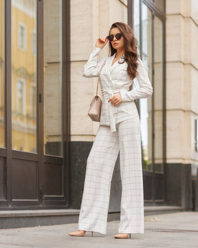 Young beautiful elegant woman in white casual costume walking at street on a summer day. Pretty lady wearing sunglasses and holding handbag. Businesswoman standing and posing in the city
