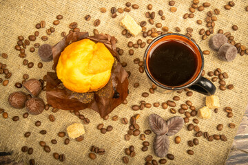 Fresh cupcake, coffee mug, chocolates and coffee beans on a homespun fabric with a rough texture. Close up.