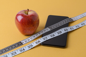 Measuring tape with centimeters and inches, smartphone and ripe red apple on a textured yellow...
