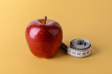 Inch measuring tape roll and ripe red apple on a textured yellow background. Concept of healthy eating, dieting, weight loss and body slim.