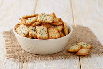 Small salty rectangular crackers with poppy and sesame seeds in a beige ceramic bowl on a white wood table. Crispy wheat flour snack and beer appetizer.