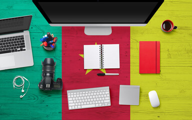 Cameroon flag background with headphone,camera, notebook and mouse on national office desk table.Top view with copy space.Flat Lay.