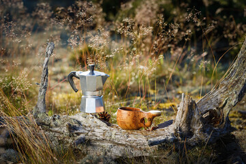 Obraz premium Coffee maker and kuksa wooden cup on tree trunk in the forest. Background is blurry.