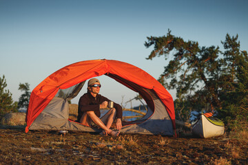 Happy man drinks tea in orange tent on island with sea kayak.