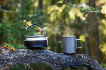 Kettle and a titanium cup of tea on a tree trunk in the forest. The background is blurred.