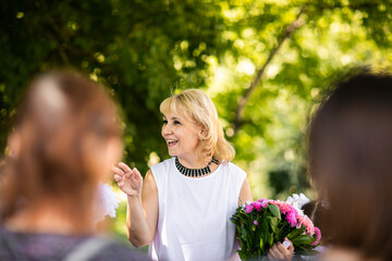 Obraz premium Adult teacher posing with bouquet of pink flowers among parents at the school yard. Green background.
