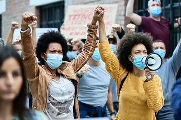 Black female activists holding hands while protesting with crowd of people during coronavirus...