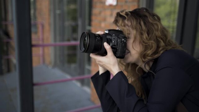 Portrait of female photographer with professional camera. Action. Side view of caucasian woman with curly hair taking pictures on her camera on red brick wall background.
