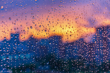 Bright fiery sunset through raindrops on window with bokeh lights. Abstract background. Water drop on the glass against the blurred silhouettes high-rise city.