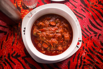 A delicious white bowl of red meat sauce or beef gravy filled with diced pieces of meat, tomatoes, onions and pepper served on a colorful black and red pattern table cloth - top view