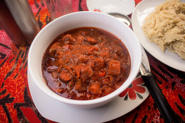 A delicious white bowl of red meat sauce or beef gravy filled with diced pieces of meat, tomatoes, onions and pepper served on a colorful black and red pattern table cloth with some mashed pasta