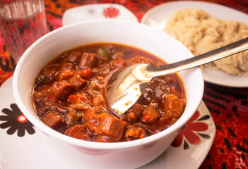 A white bowl of red meat sauce or beef gravy filled with diced pieces of meat, tomatoes, onions and pepper served on a colorful black and red pattern table cloth with a silver spoon and mashed pasta