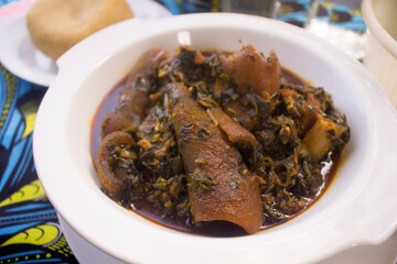 A white bowl of tasty Nigerian vegetable soup cooked with assorted meat, dried fish, cow skin (ponmo or kpomo or pomo) and Garri or Eba. Served on a colorful blue African pattern table cloth