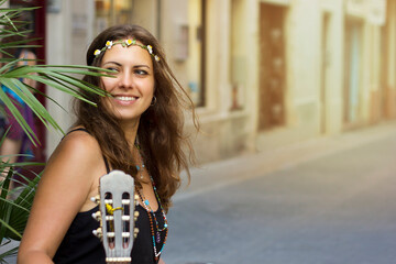 Fototapeta premium beautiful happy young girl with curly hair hippie style with a guitar and a wreath on her head next to a palm branch. Woman photo portrait waist-high on the street. Free lifestile. Street musicians