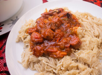 A white plate of tasty mashed pasta served with some meat sauce or red spicy beef gravy containing diced pieces of meat, sausages, sliced onions, tomatoes and pepper