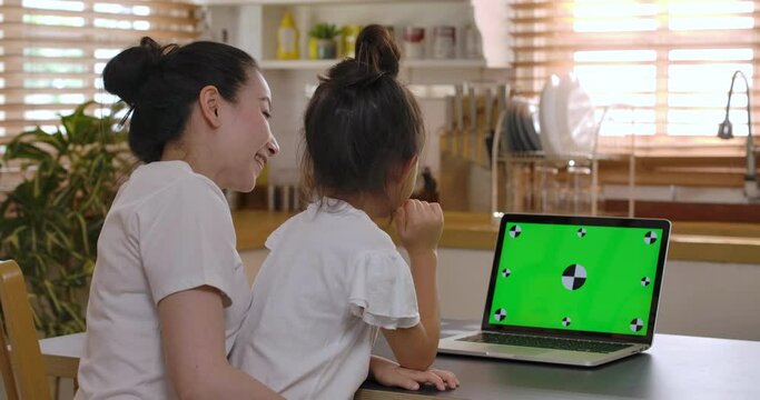 Happy young asian mother and cute little kid daughter using laptop computer with green screen talking with friends or family in webcam conference chat application. Family videocall concept.