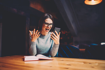 Emotional astonished young woman reading shocking information from internet sources on mobile phone device sitting in university.Hipster girl watching disgusted video received from friends on cellular