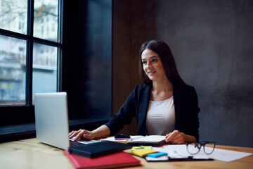 Intelligent female economist dressed in elegant formal wear working in office with online documents in database preparing for quarterly report on conference meeting with executive managers using wifi