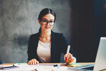 .Young professional designer in stylish eyeglasses holding marker in hand and working on developing new graphic sitting in stylish studio at desktop with sketch and modern laptop computer device.